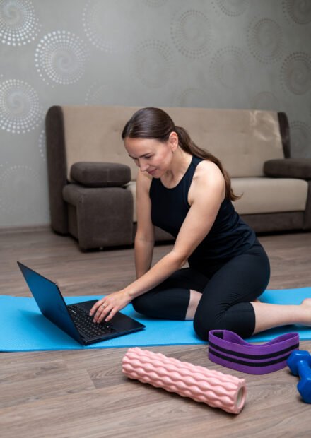 Young woman exercising at home with dumbbells, foam roller, resistance band and yoga mat, following online fitness program, healthy lifestyle