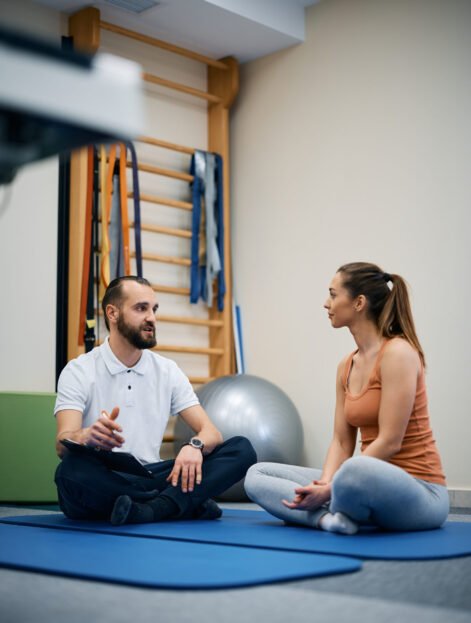 Physical therapist and athletic woman talking while sitting on the floor at health club. Copy space.
