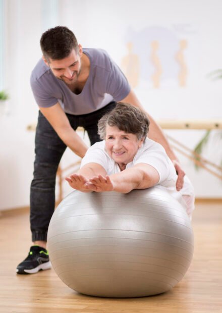 Smiling physiotherapy student helping senior woman lay on the exercising ball during rehabilitation