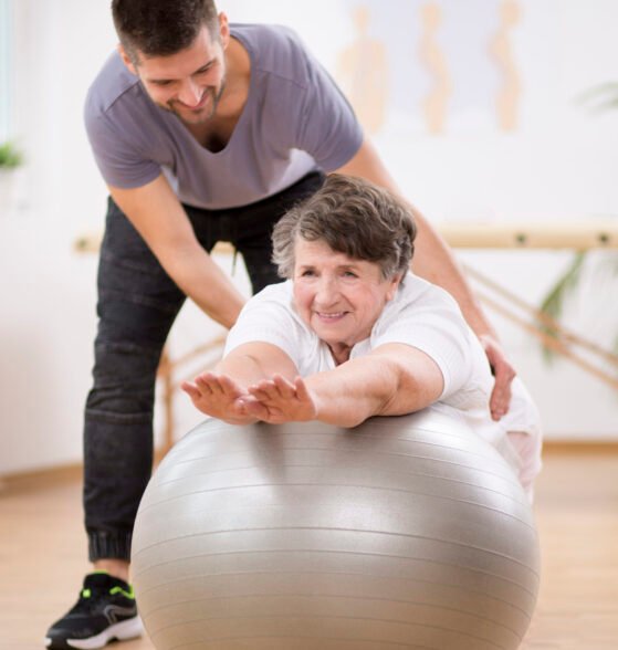 Smiling physiotherapy student helping senior woman lay on the exercising ball during rehabilitation